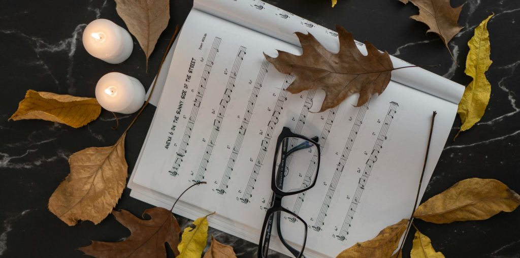 Cozy autumn setup with sheet music, glasses, and candles surrounded by leaves on a marble surface.